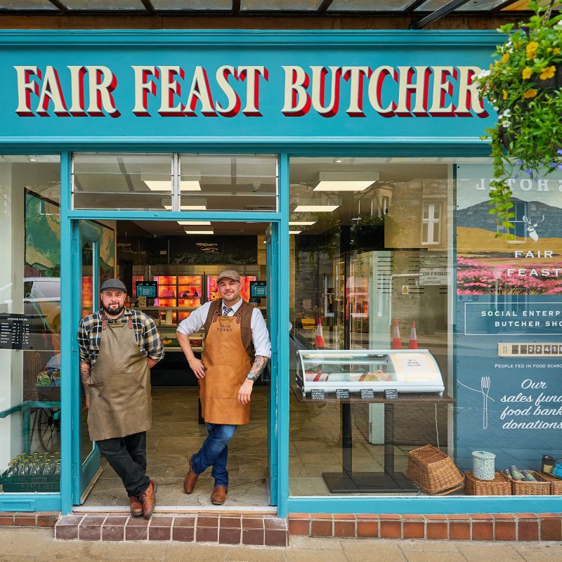 Two smiling men wearing aprons which say 'Fair Feast' on them standing in front of the Butchers shop.