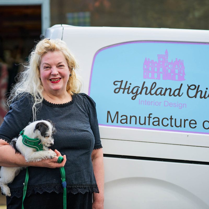 A smiling woman holding a small black and white dog standing in front of a van for Highland Chic.