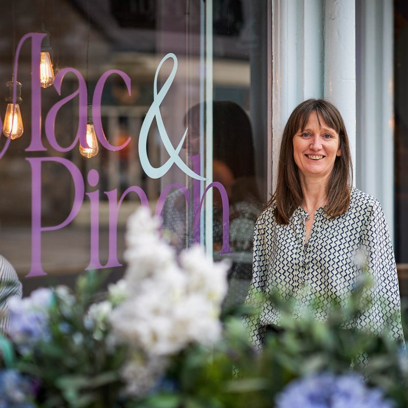 A smiling woman standing in front of a window with a purple decal on it which says 'Mac & Pinch'