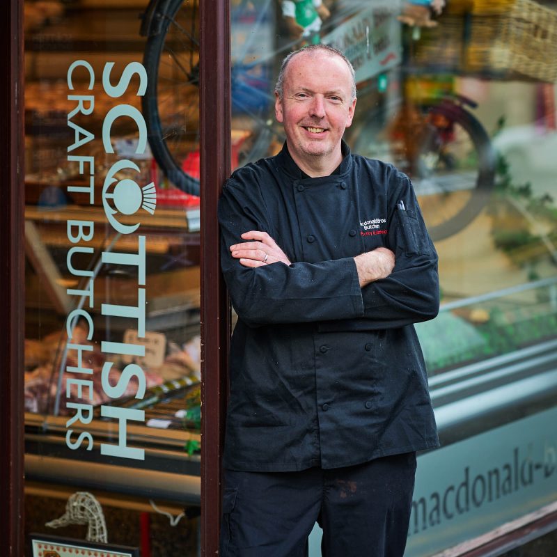 A smiling man wearing a black chefs jacket standing in front of the butchers shop.
