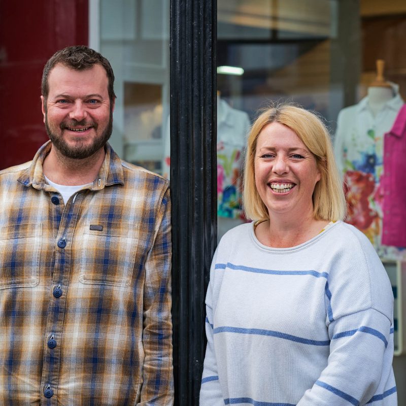Two smiling people standing outside the shop leaning against a black pole.