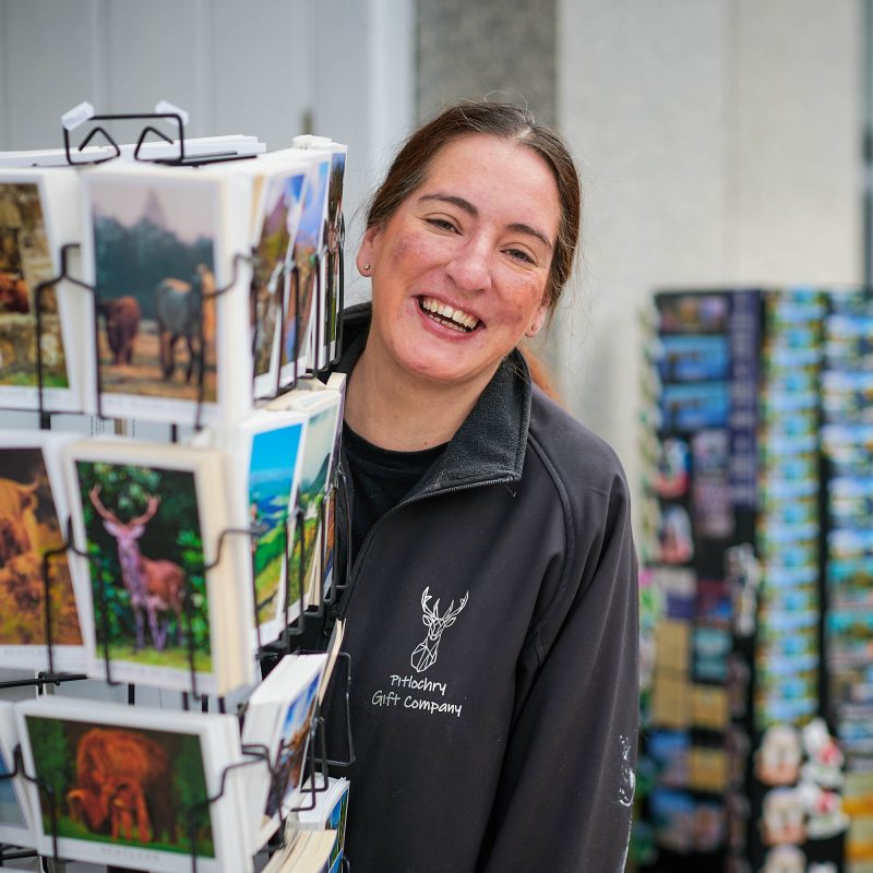 A smiling woman wearing a fleece which says 'Pitlochry Gift Company' on it. She is standing behind a postcard stand.