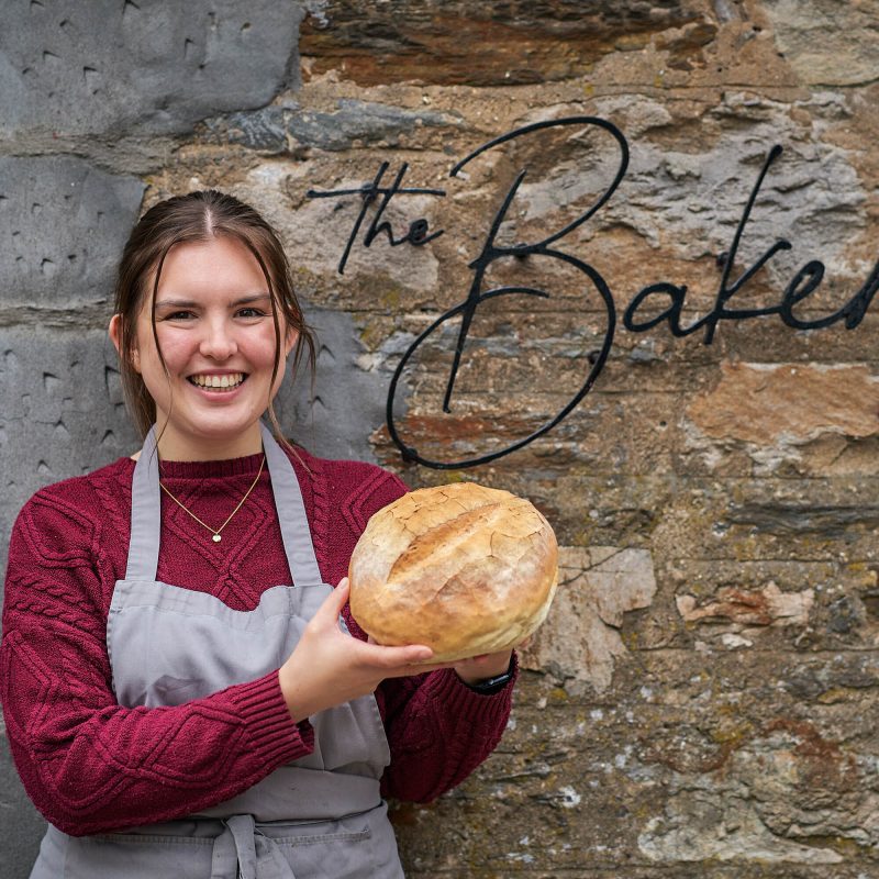 A smiling woman holding a loaf of bread standing next to a sign which says 'The Bakery'