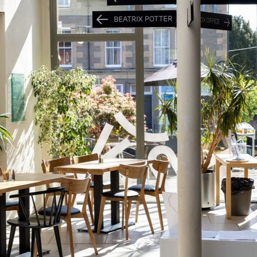 Room with large windows, a sunny day, wooden sets of tables and chairs in foreground, potted plants in background. column displaying signage for Beatrix Potter garden and box office