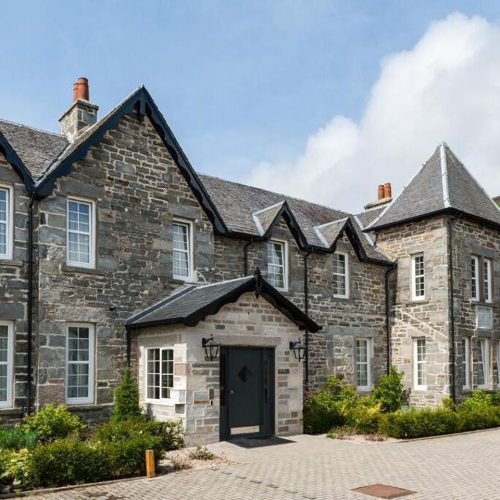 Large old stone building with black painted trim, green hedging under a blue sky