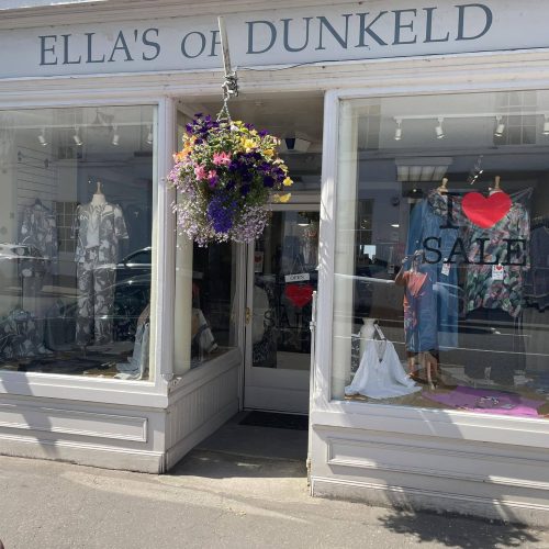 Cream painted shop front, displaying ladies clothes, brightly coloured flower hanging basket above doorway and sign that says Ellas of Dunkeld