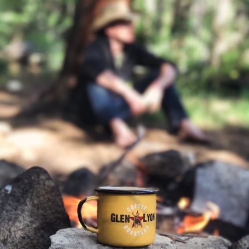Metal coffee cup in foreground with logo of Glen Lyon Coffee Roasters on it , man wearing a hat sits under a tree on a sunny day in background.