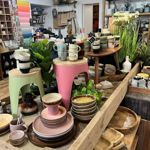 Coloured crockery, pink and yellow stools, plants, cups on display on a large wooden table with brightly coloured  racks of wrapping paper on wall behind