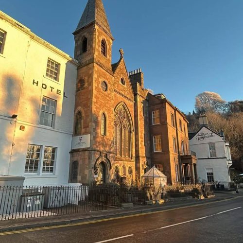 Large old church with tall spire located between white building on a sunny day, with blue skies, sign on side of church says Jeffreys Dunkeld