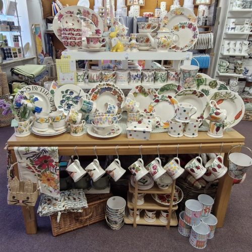 Wooden display table with brightly coloured mugs, plates, tea towels, bowls in foreground with more items on walls behind