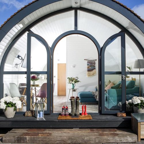 View of a glamping pod with doors open and glass frontage,  internal view of sofa and dining set, pots of flowers externally and wellie boots on doormat.