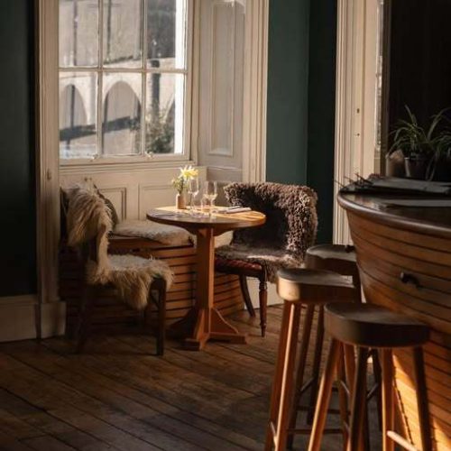 Sunlight streams in a Victorian window onto a wooden floor with a bar stools and a cosy set of chairs and table in front of window