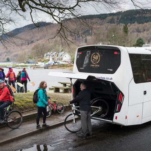 Man removing a bike from the back of a large white bus, with people on bikes in front of a loch with green hills in background.