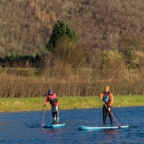 Two men on blue paddleboards on water with hills and greenery behind them.