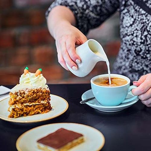 Woman pouring milk into a coffee cup on a table with cakes and chocolate treats