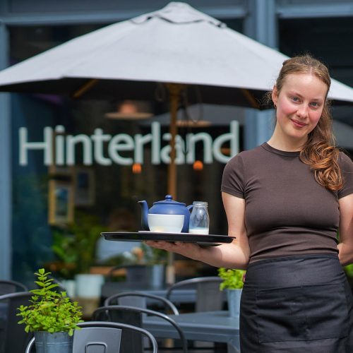 A smiling woman wearing a brown t-shirt and black appron. She is holding a tray with a navy teapot, a white cup and small jug of milk.