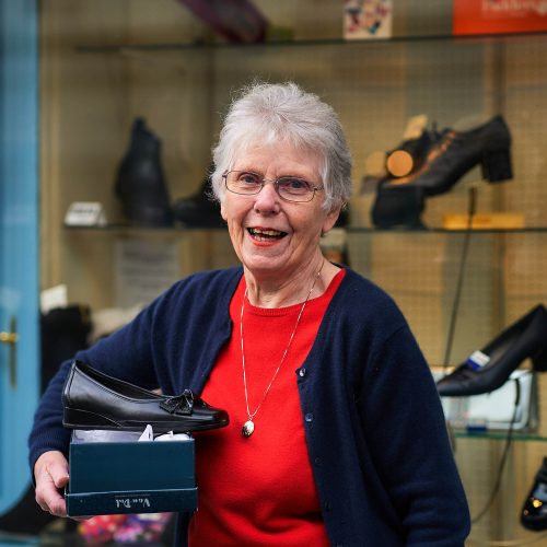 Smiling grey haired lady holding a box with shoes, in front of a  shop window with blue trim displaying shoes.