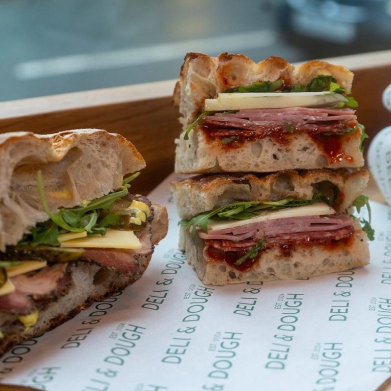Two deli style sandwiches with lots of filling, sitting on a shelf in front of a window