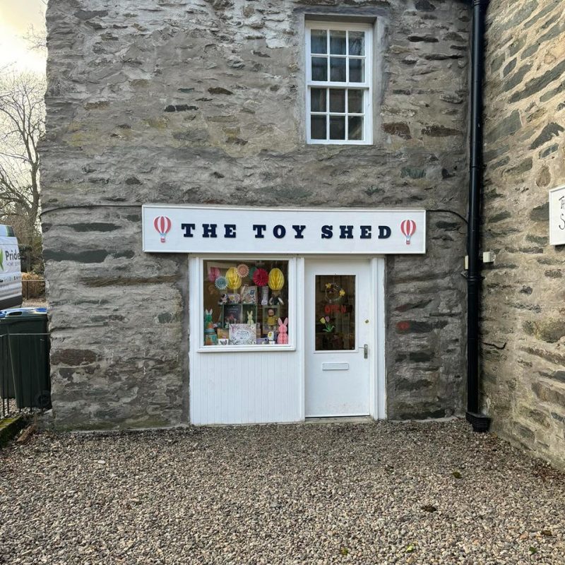 Old stone building, with white door and window with bunnies and children's items on display. Sign says The Toy Shed with hot air balloons either side.