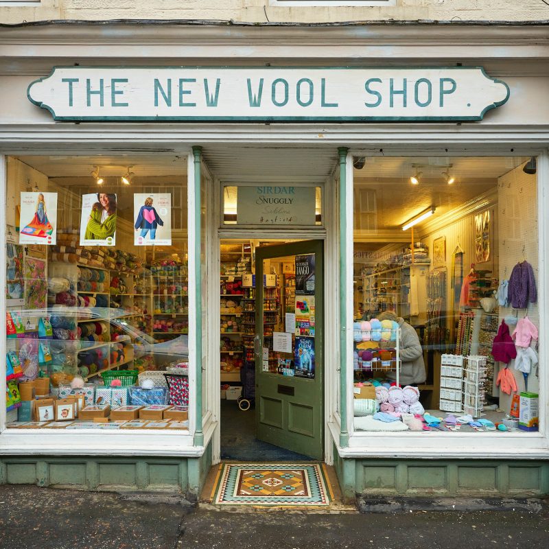 Shop front with large windows displaying knitted goods and wool in bright colours, sign says The New Wool Shop