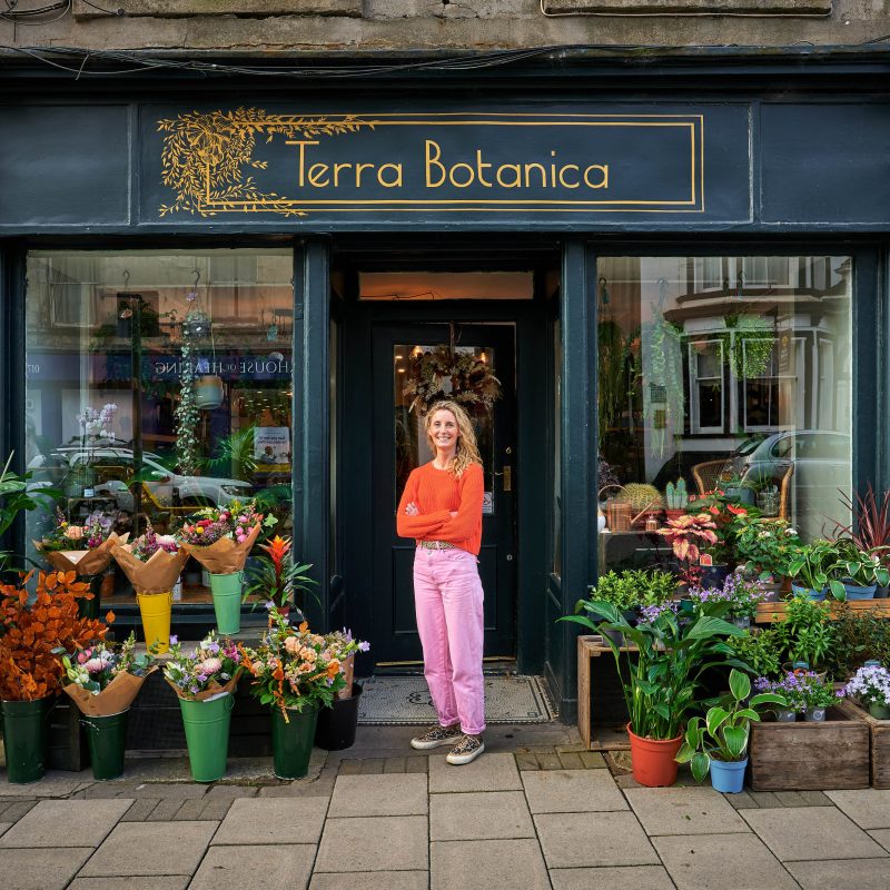 Smiling blonde haired lady wearing an orange jumper and pink trousers, outside a shop displaying flowers and plants sign says Terra Botanica
