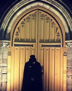 Figure wearing black cape and top hat standing in front of a large wooden Church door