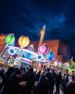 Brightly coloured  funfair lights on a ride, with people in foreground, under a dark nightime sky.