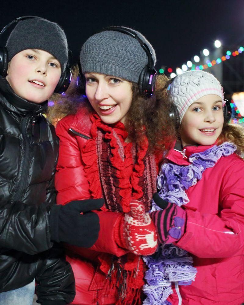 Three smiling people wearing hats and cosy jackets, with headphones on, outside with christmas lights in distance.