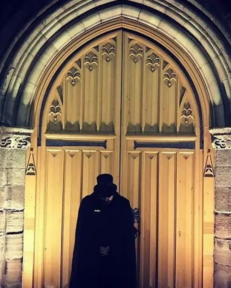Figure wearing black cape and top hat standing in front of a large wooden Church door