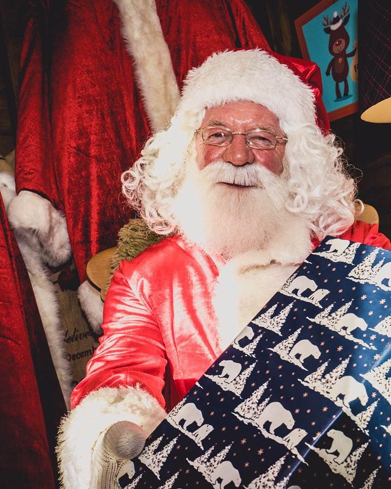 Smiling Santa with glasses and white beard holding a large blue Christmas present