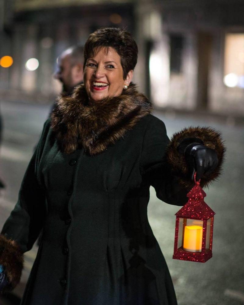 Smiling dark haired woman ,dressed in wintercoat and cosy scarf, holding a red lantern with a lit candle inside.