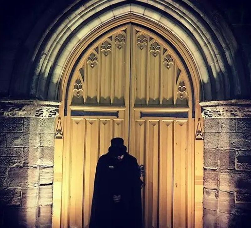 Figure wearing black cape and top hat standing in front of a large wooden Church door