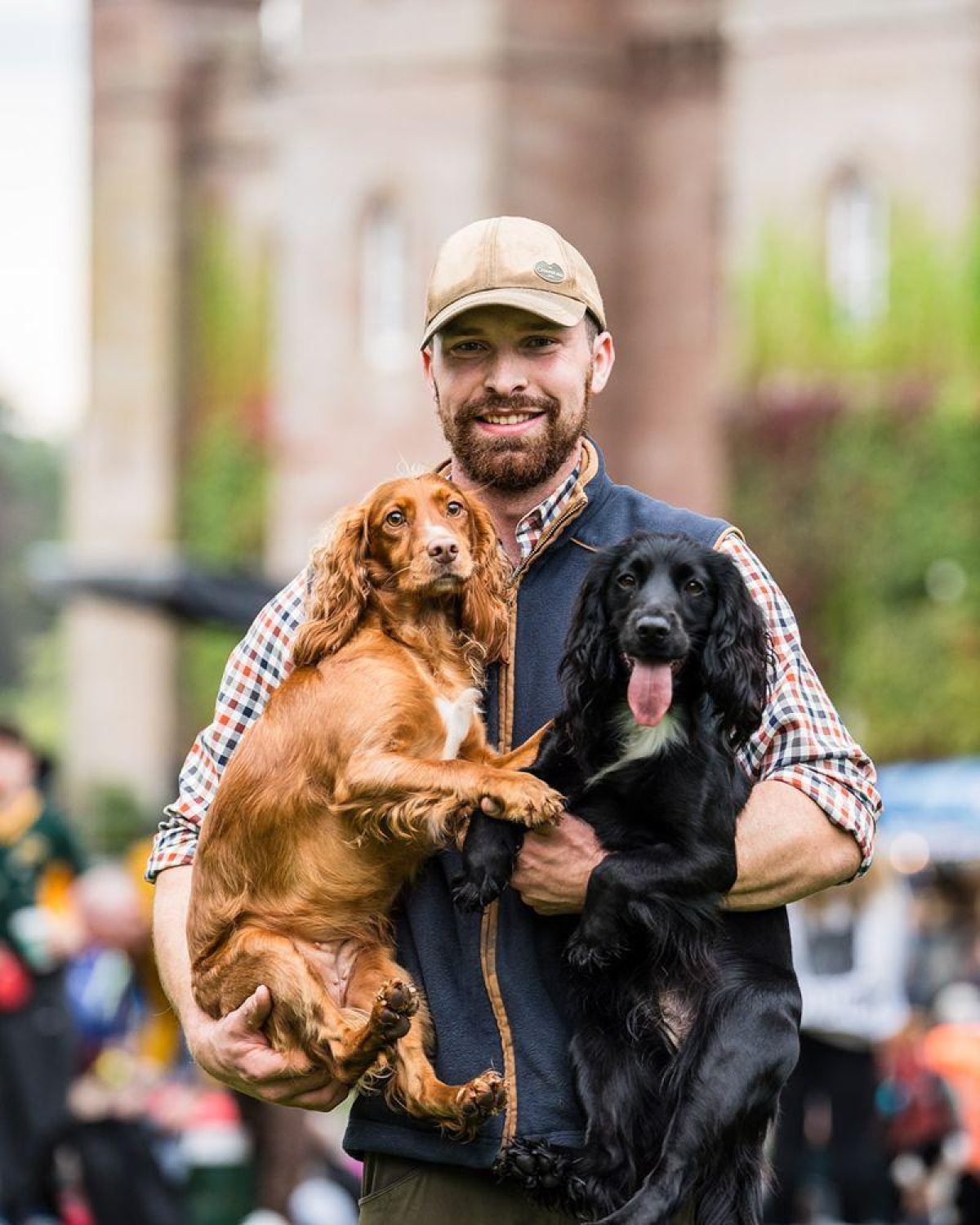 Smiling bearded man in a baseball hat carrying two dogs in front of Scone Palace