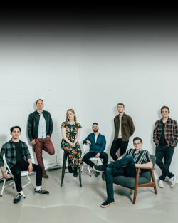 A group of people posing in front of a white screen. Some are standing and some are sitting on chairs and stools.