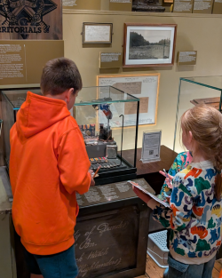 A boy wearing a neon orange jumper and a little girl wearing a floral jumper inspecting objects in the museum.