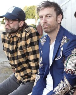 Man in yellow and black plaid shirt ,wearing glasses with a baseball cap beside a man in a blue blazer with ornate designs, both seated