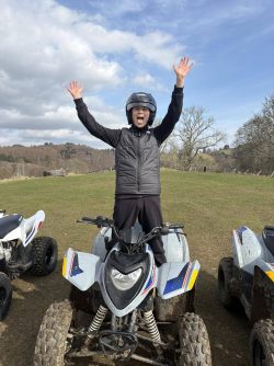 An excited boy standing on a quad bike wearing a helmat and protective gear waving his hands around.