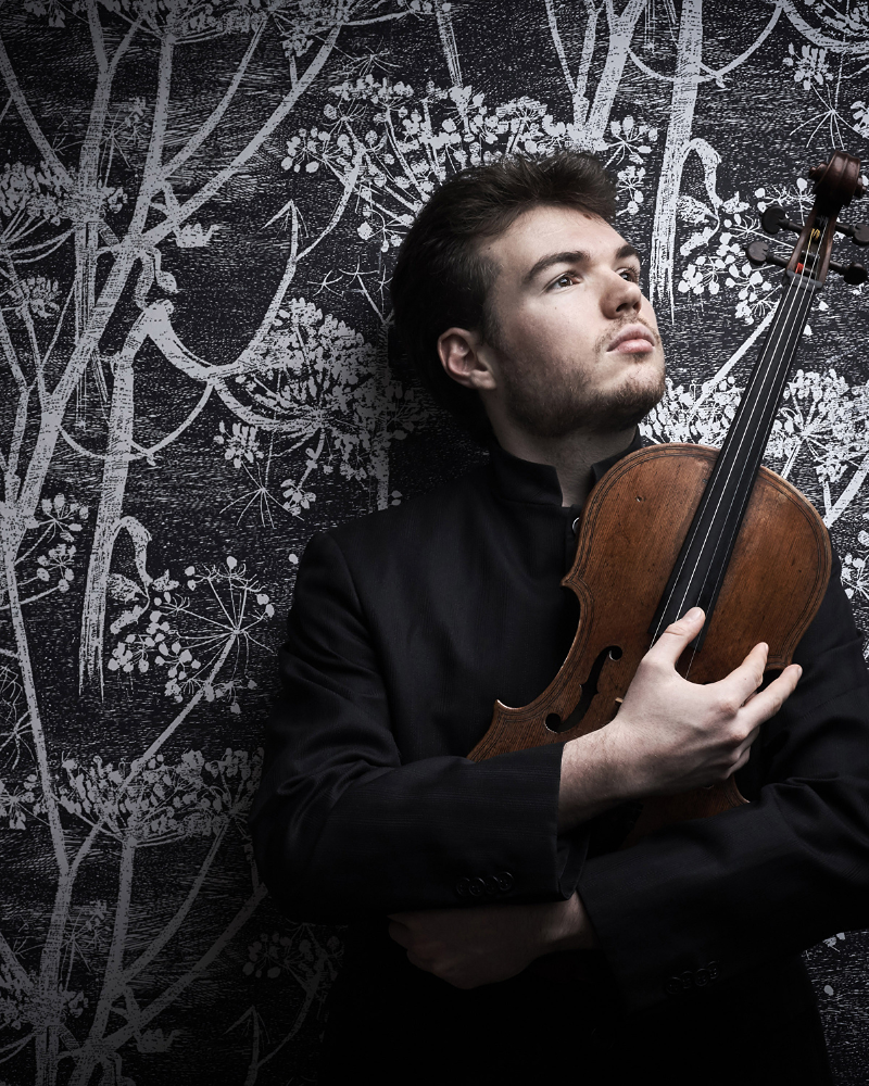 A man wearing all black holding a violin looking longingly into the distance.