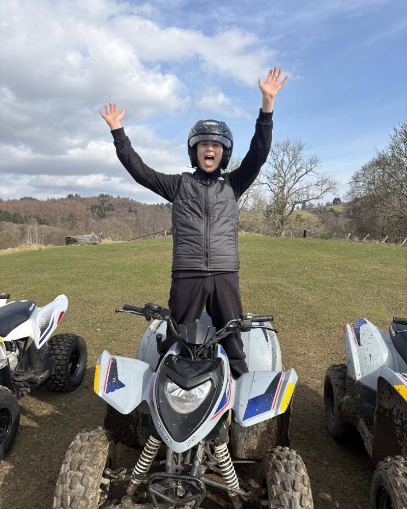 An excited boy standing on a quad bike wearing a helmat and protective gear waving his hands around.