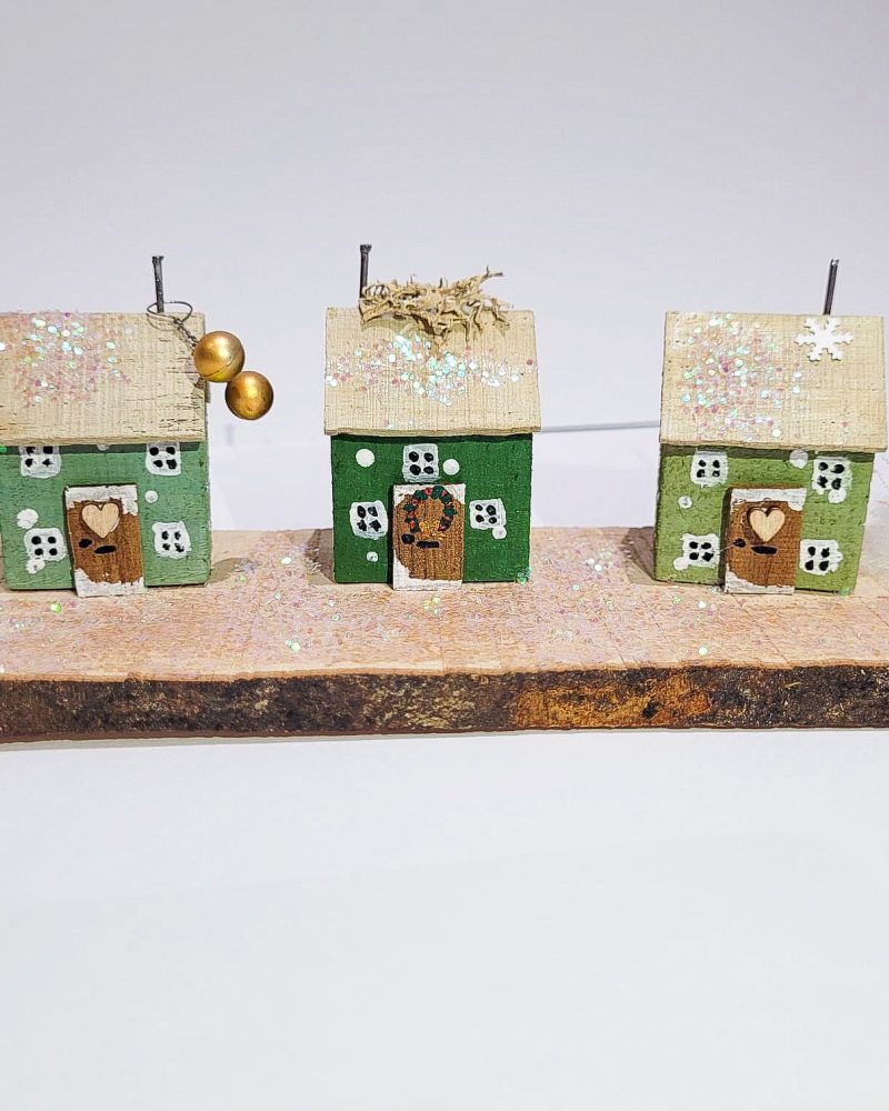 3 handmade green wooden cottages along a plank of wood covered in fake snow.