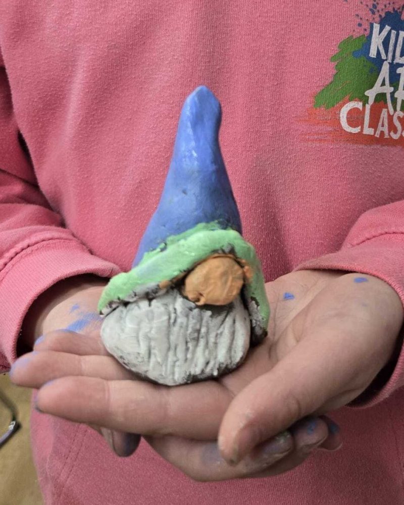 A child in a pink hoodie holding a handmade gonk with a blue and green hat made from clay.