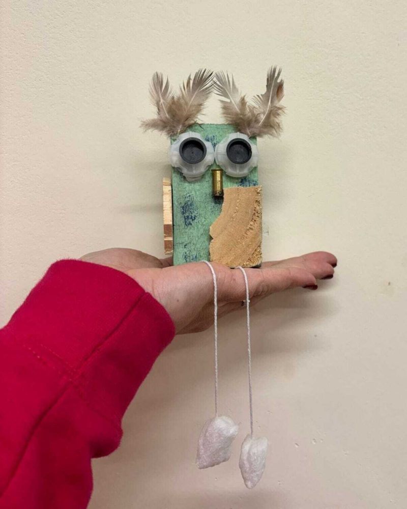 A handmade green owl with light wood wings and feathers being held up by a hand.