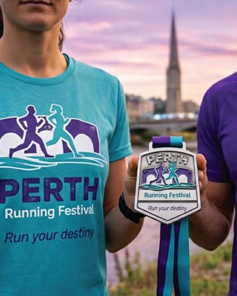 Two people holding a medal, and wearing t-shirts that say Perth Running Festival with view of Perth in background