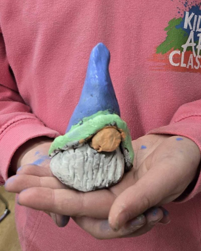 A child in a pink hoodie holding a handmade gonk with a blue and green hat made from clay.