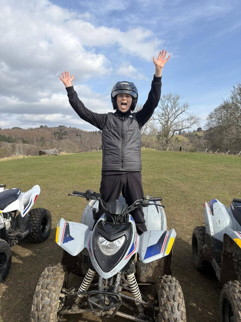 An excited boy standing on a quad bike wearing a helmat and protective gear waving his hands around.