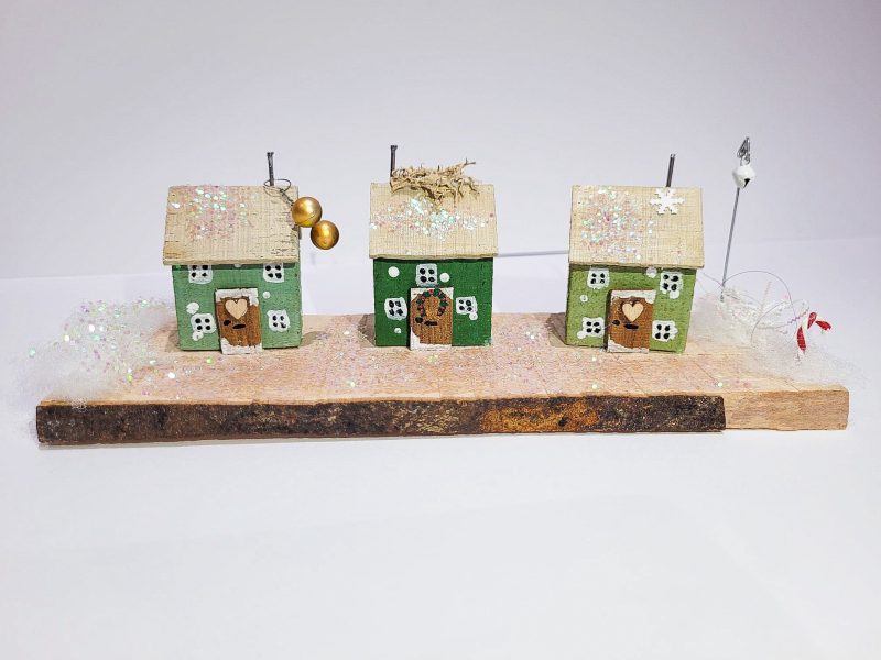 3 handmade green wooden cottages along a plank of wood covered in fake snow.
