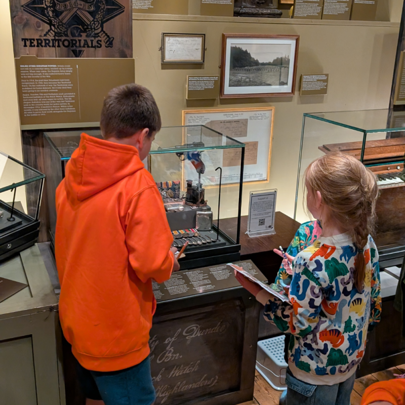 A boy wearing a neon orange jumper and a little girl wearing a floral jumper inspecting objects in the museum.