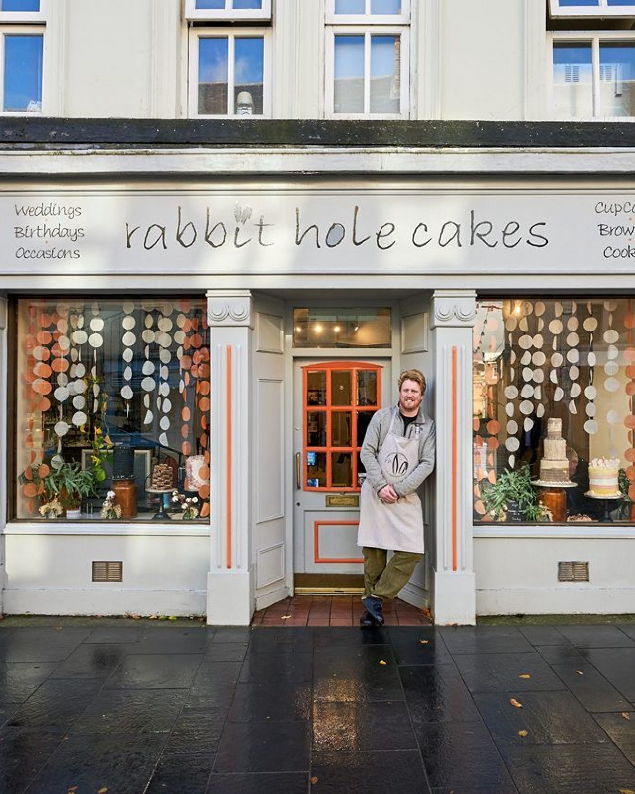 A man leans against a window displaying tiered cakes