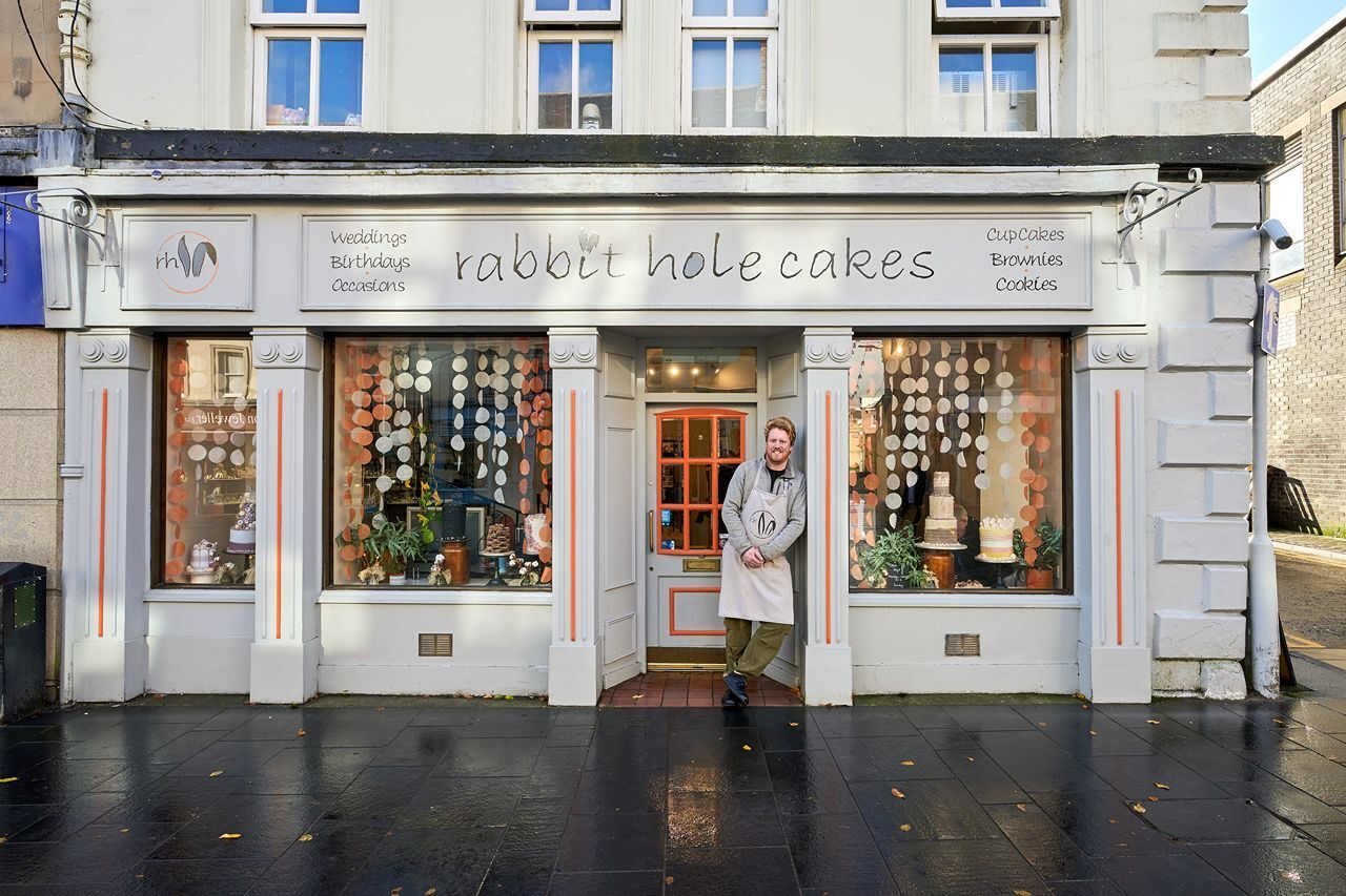 A man leans against a window displaying tiered cakes