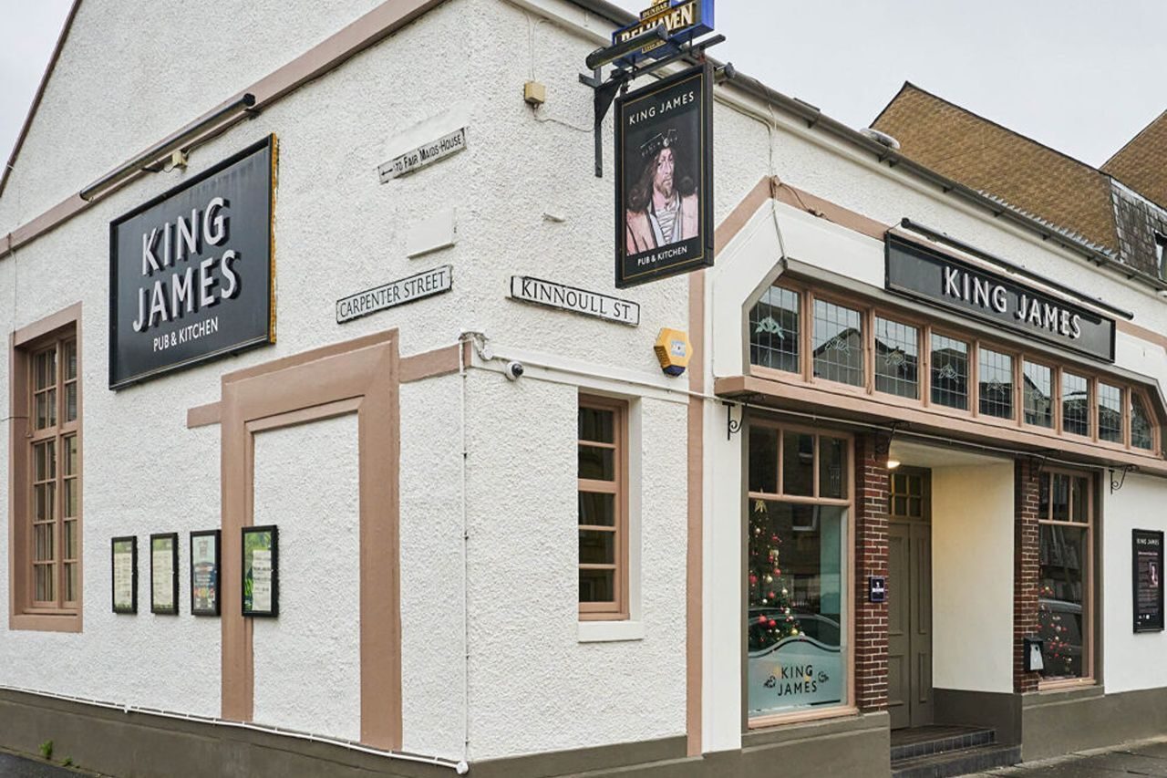 A white pub building in Perth with beige windows and a black sign with a portrait of Scottish King James